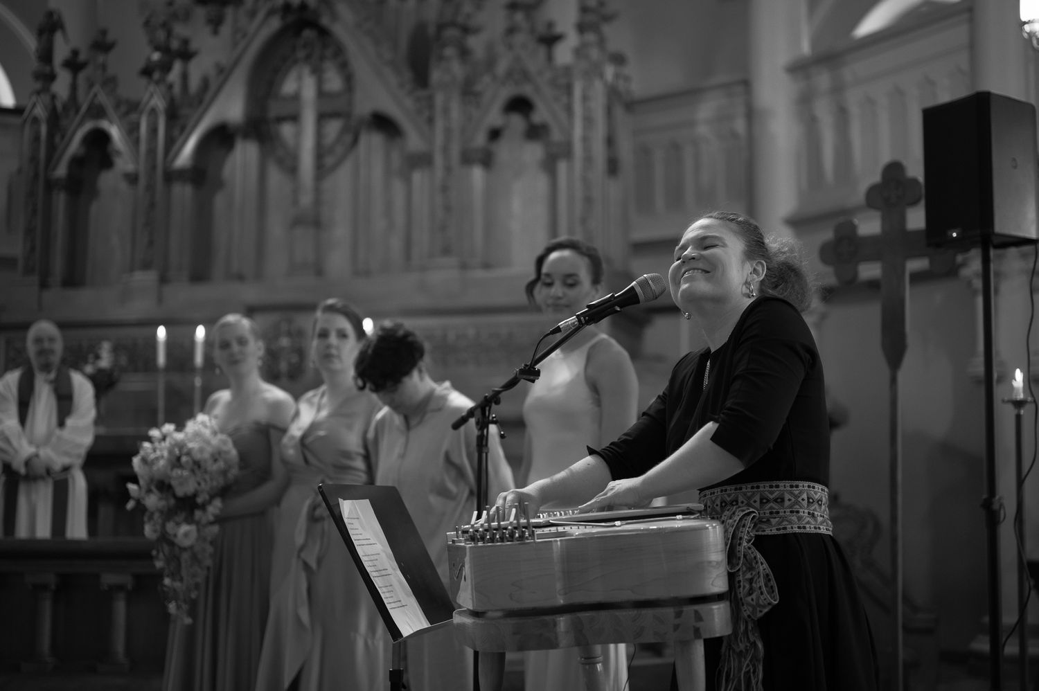 Black and white photo of a woman singing at a church wedding ceremony with bridesmaids and clergy in the background.