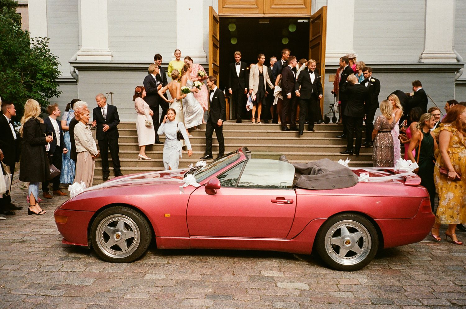 Red Porsche 968 convertible parked outside a wedding venue with guests gathered on the steps.