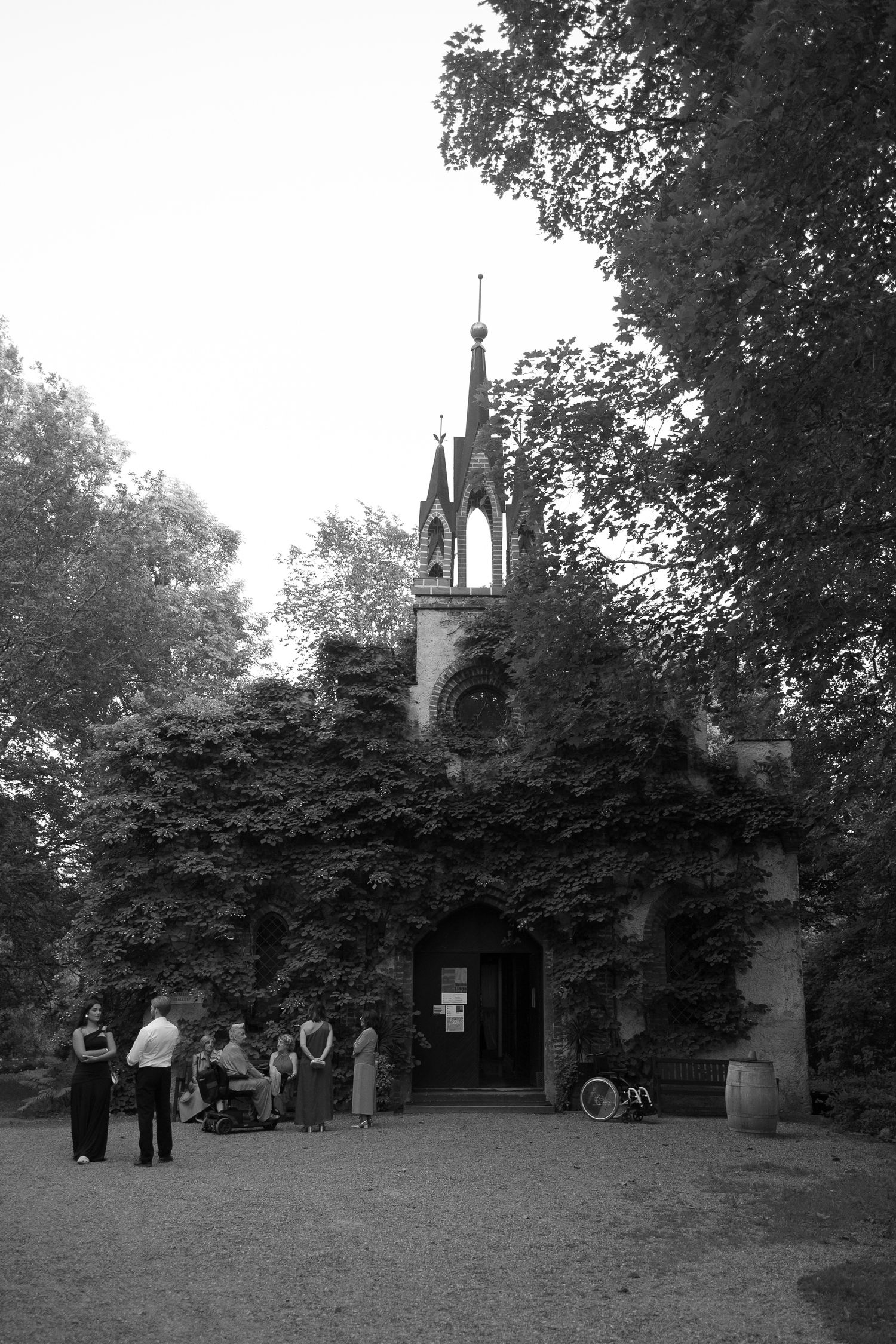 Black and white photo of people gathered outside an ivy-covered Gothic chapel with a spire, surrounded by trees.
