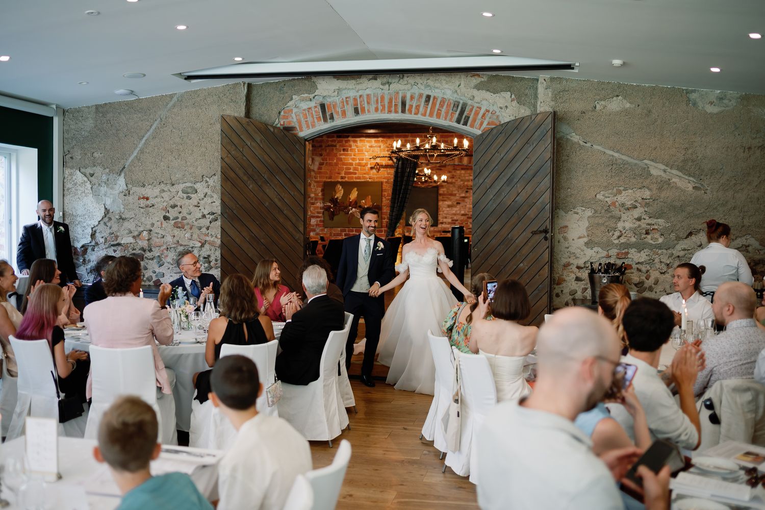 Bride and groom make grand entrance into elegant stone-walled wedding reception hall with seated guests applauding.