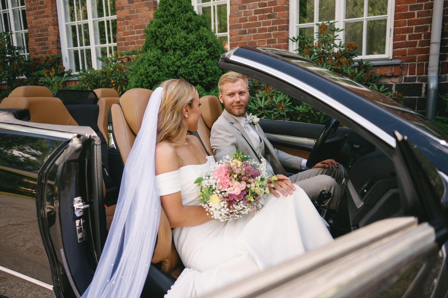 Wedding party helping bride with flowing dress enter black vehicle.