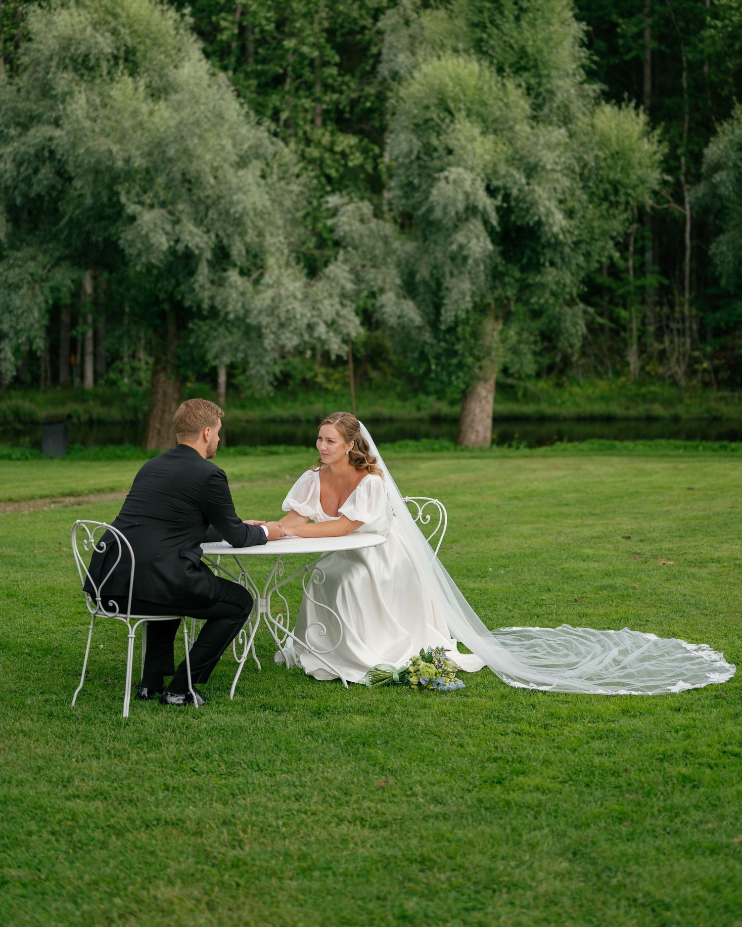Bride and groom relaxing on white garden furniture on a lush green lawn.