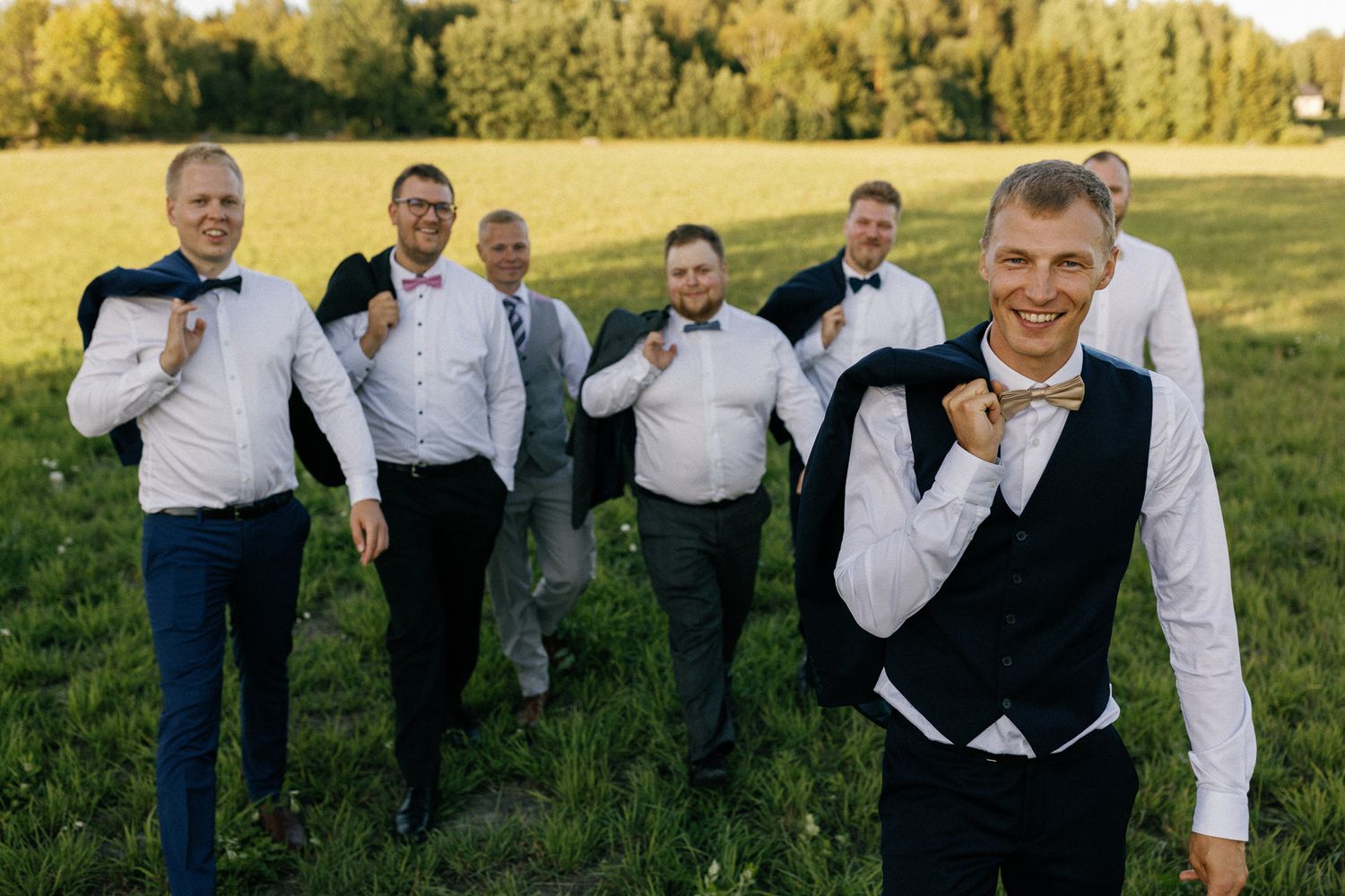 Groomsmen in suits playfully pose together outdoors in a sunny field.