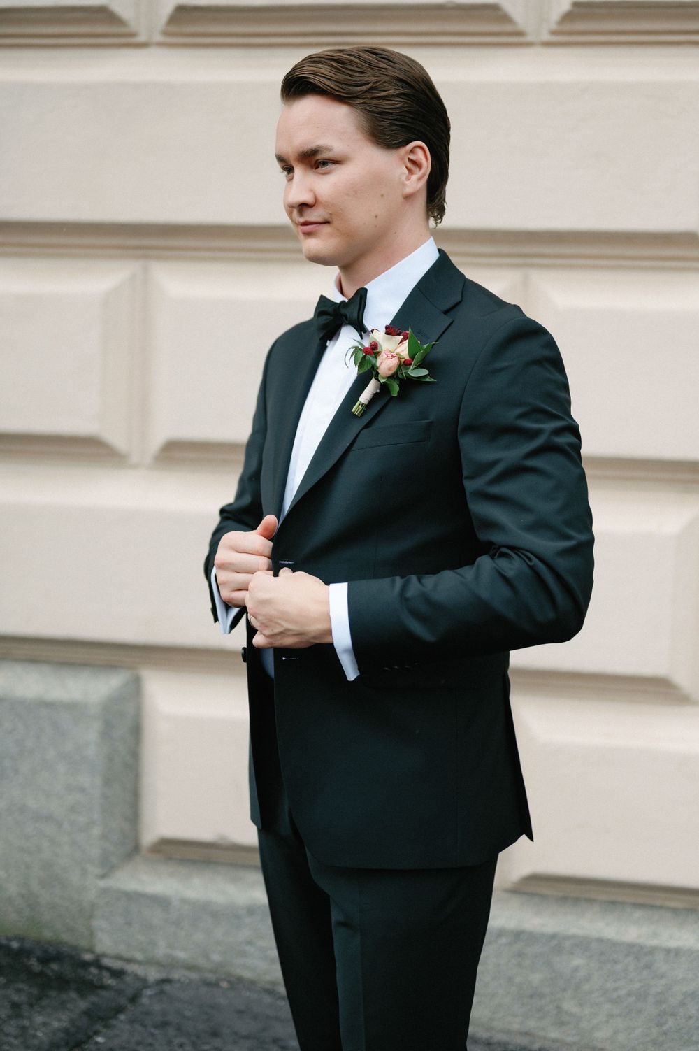 Groom in classic black tuxedo adjusts boutonniere while standing against light colored building.
