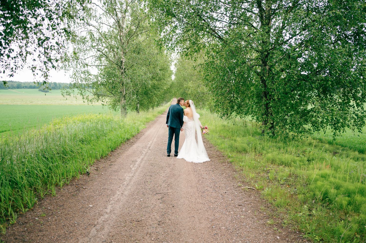 Newlyweds walking hand in hand down a scenic rural path lined with birch trees.
