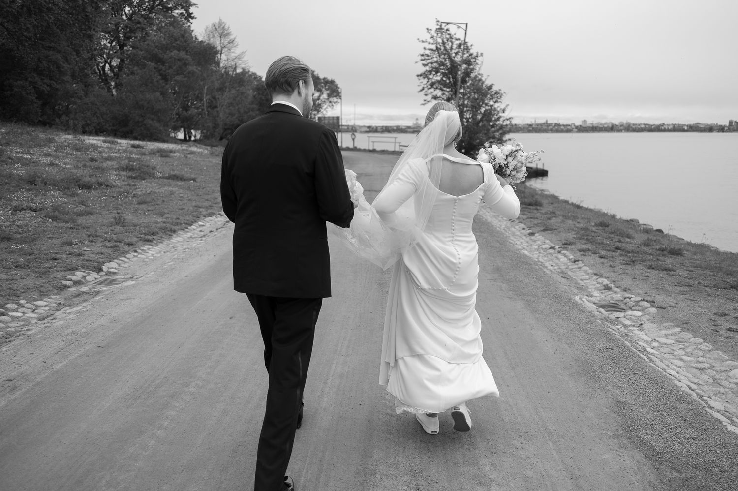 A couple walks along a scenic waterfront path in formal attire, captured in black and white photography.