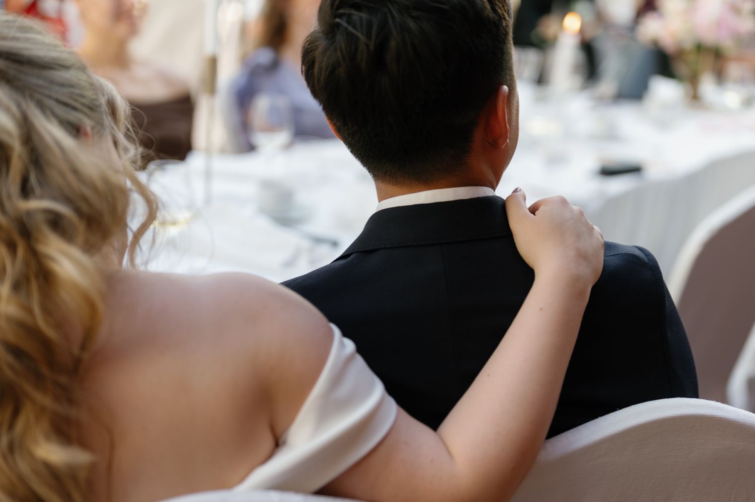 Bride and groom sitting together at wedding reception, viewed from behind, bride's hand resting on groom's shoulder.