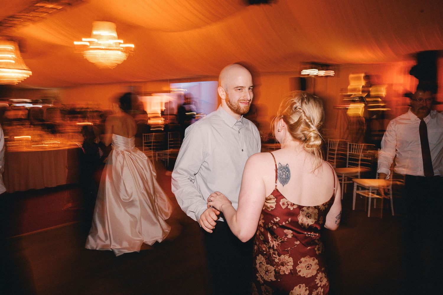 Couple dancing together at a wedding reception in a warmly lit venue with other guests in the background.
