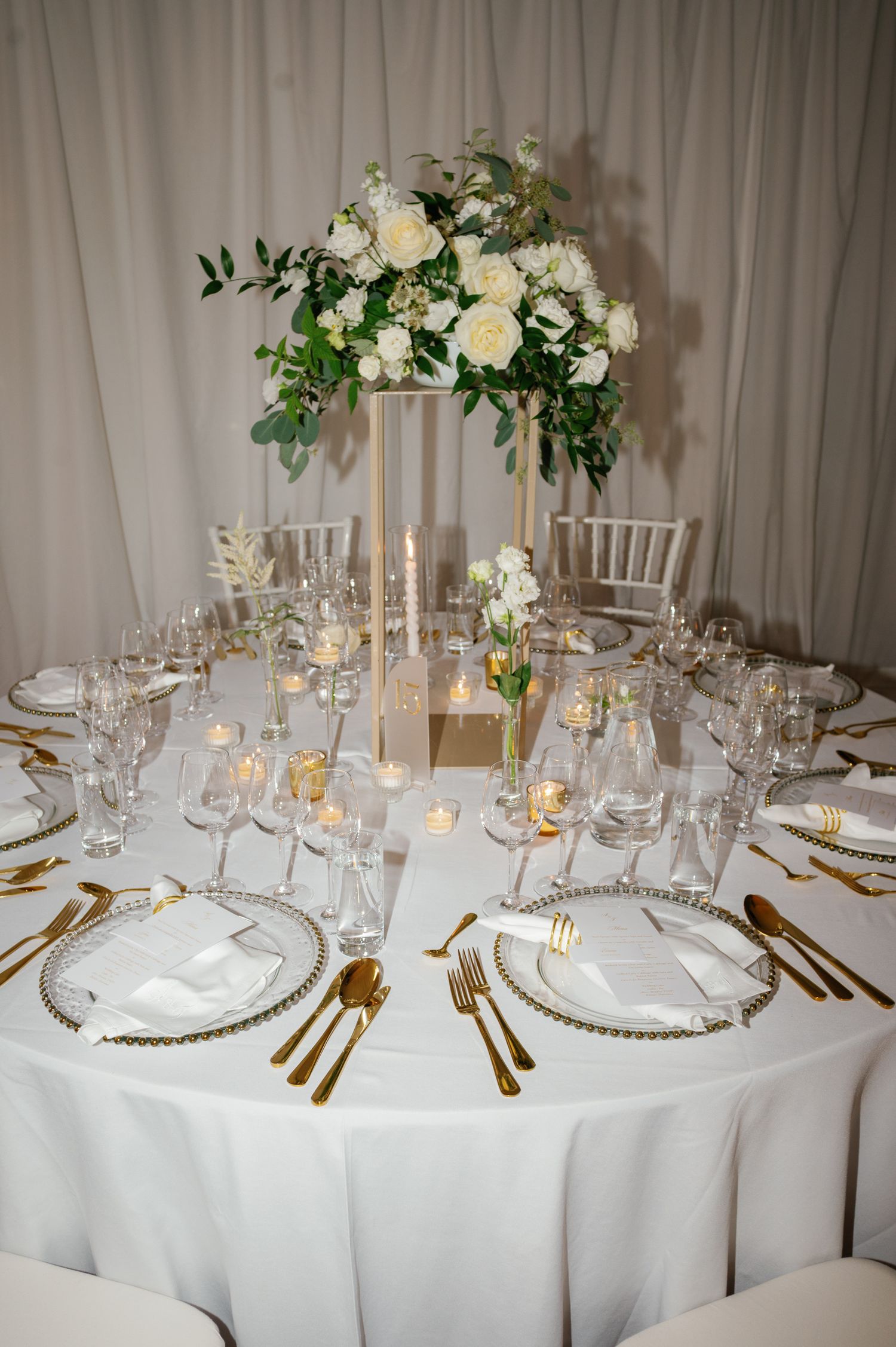 Elegant wedding reception table with white floral centerpiece, gold cutlery, crystal glasses, and white linen tablecloth.