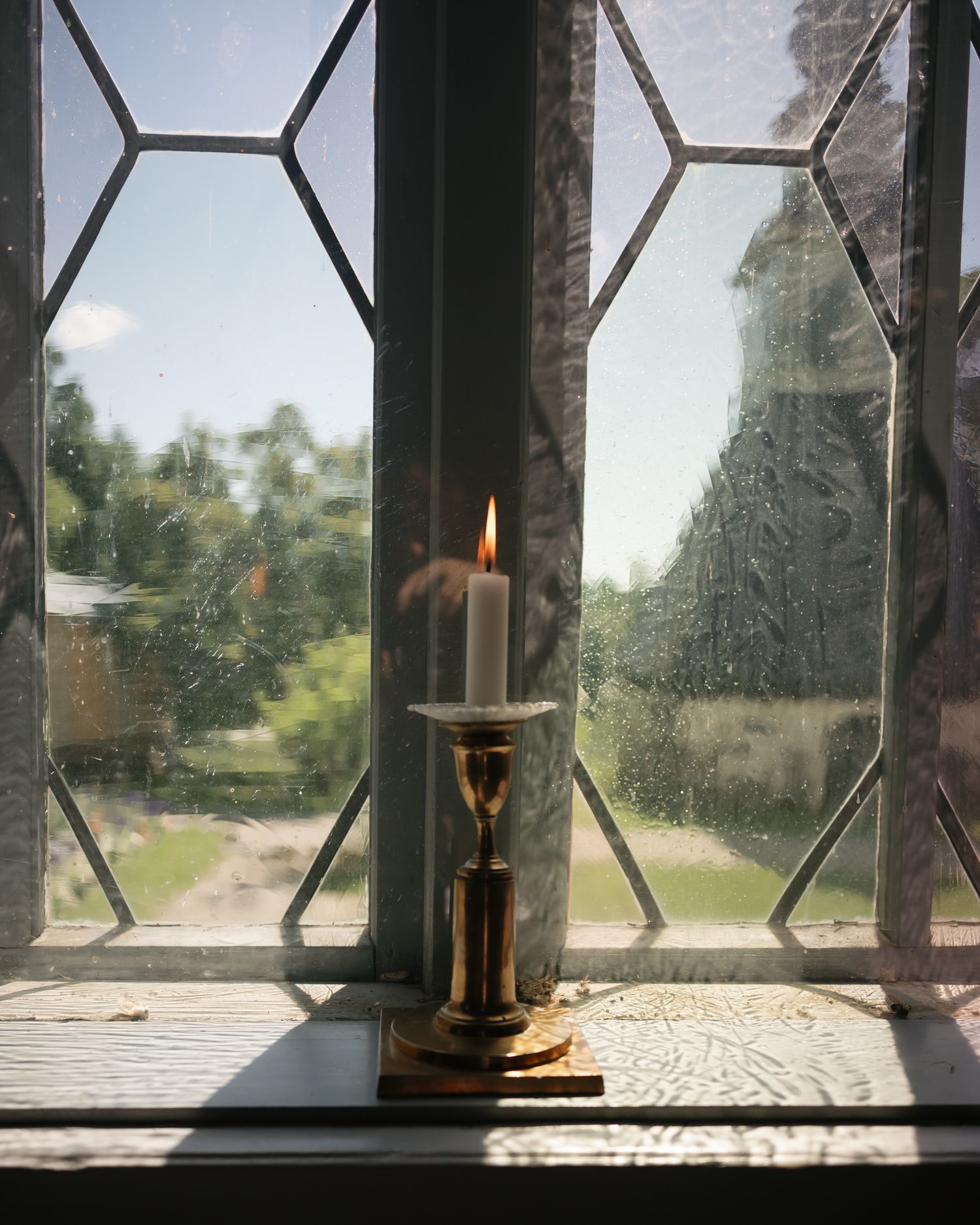 A lit white candle on a brass candlestick holder sits on a windowsill with leaded glass windows on a rainy day.