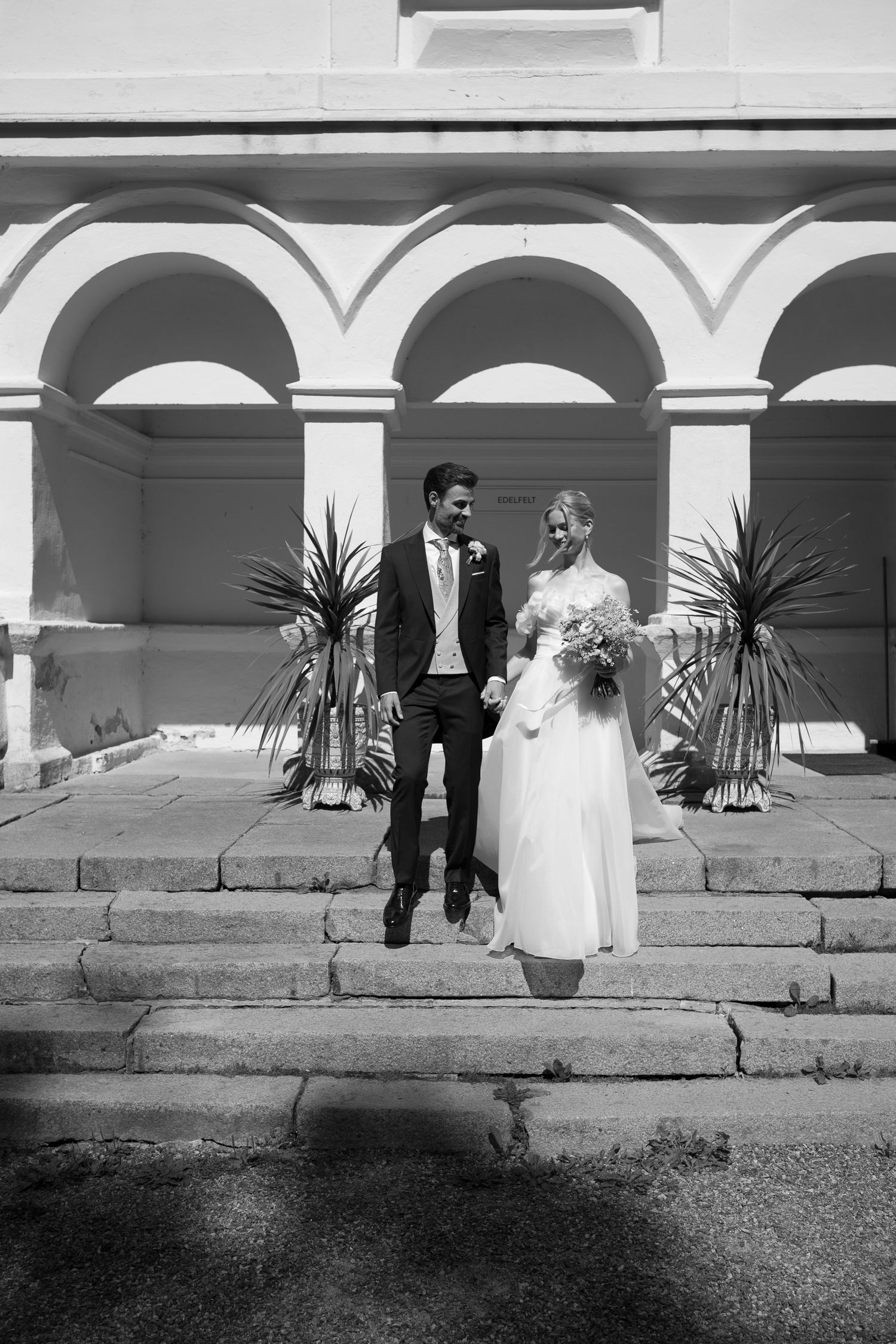 Black and white photo of a bride and groom holding hands on stone steps outside a building with arched columns.