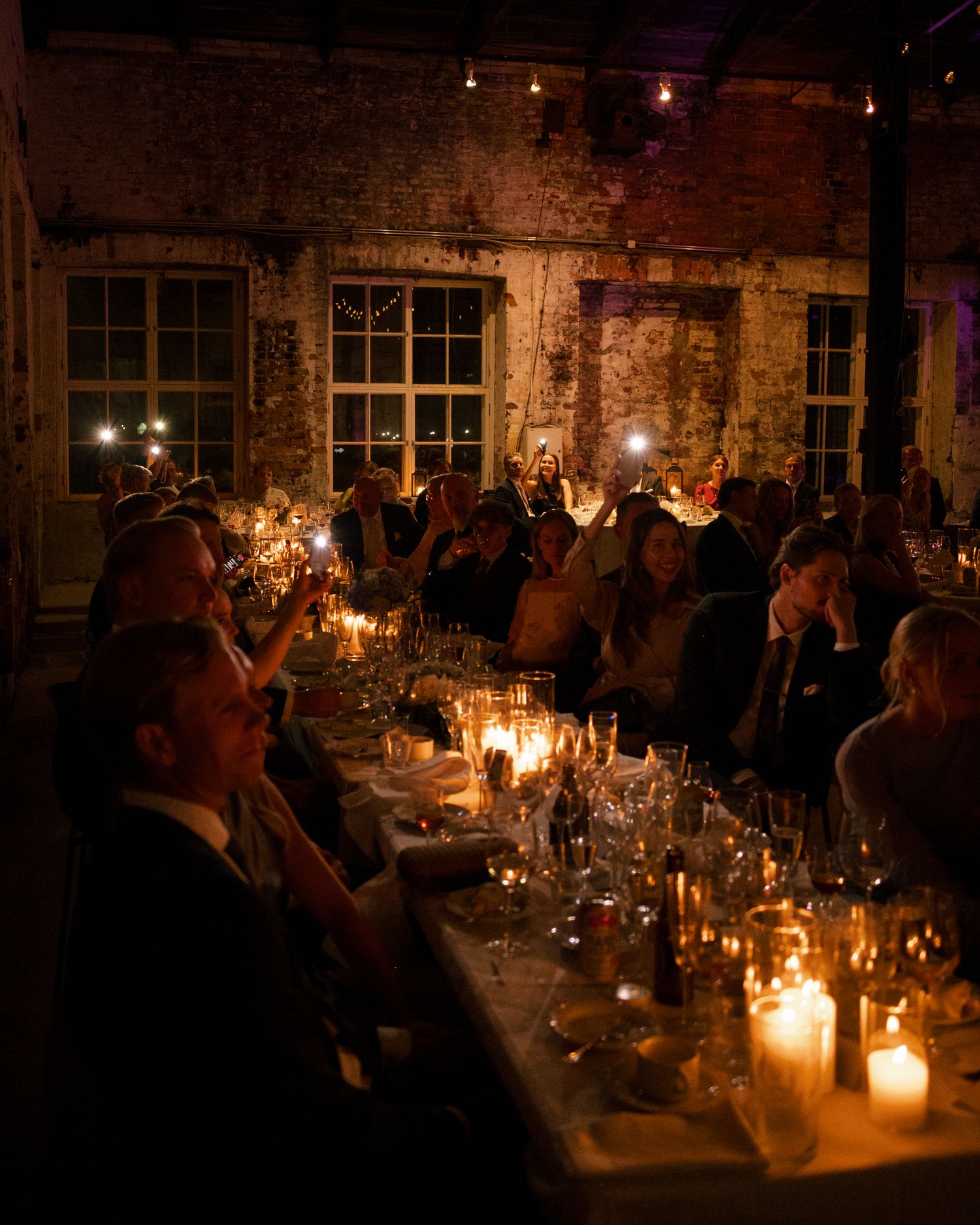 Guests seated along a long candlelit table in a rustic brick venue with large windows, enjoying an intimate evening dinner celebration.