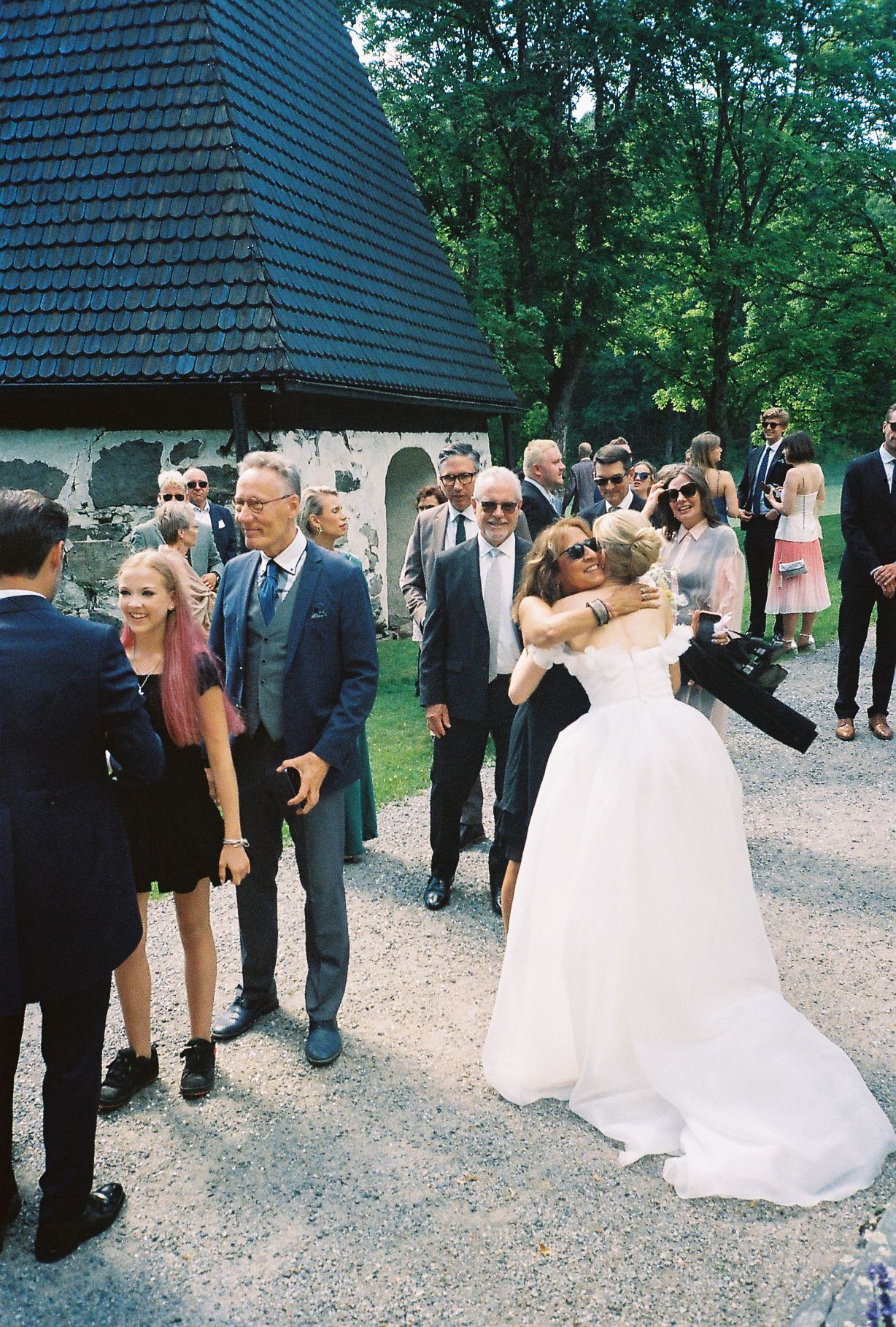 Bride in white gown hugging guest outside historic stone chapel at outdoor wedding celebration with guests gathered nearby.