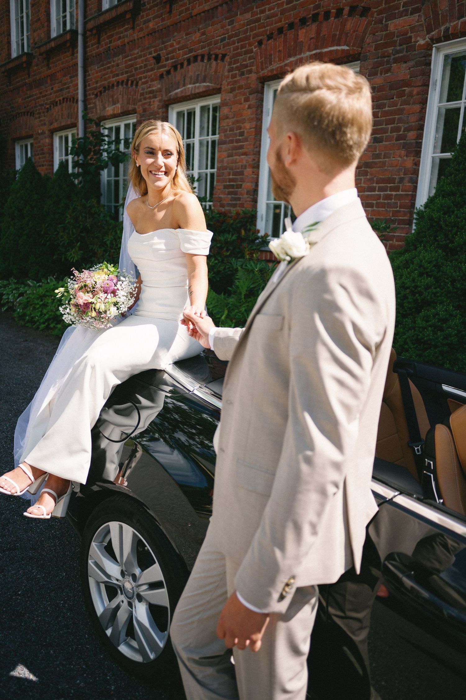 Bride in white dress being helped into a black car outside a brick building.