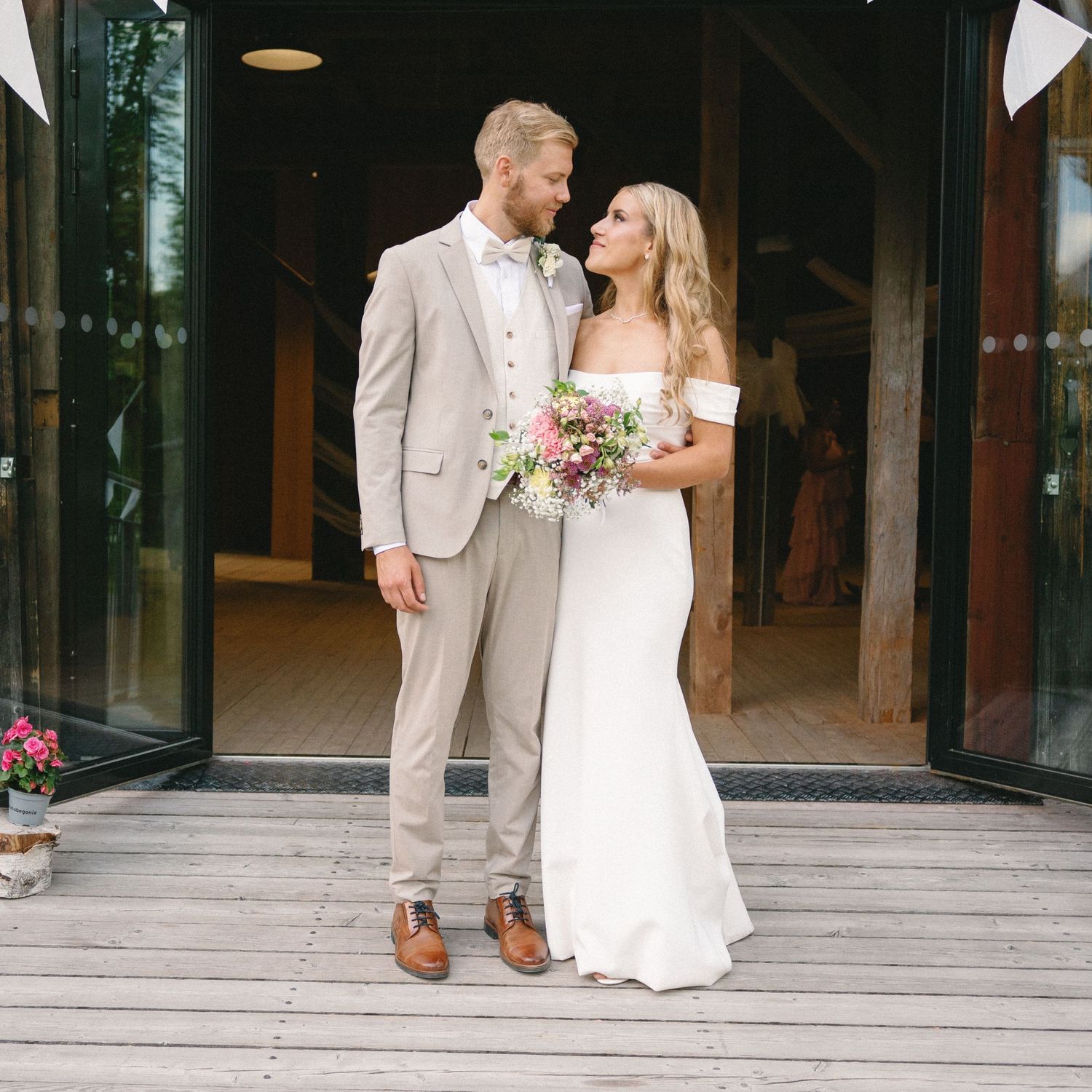 A couple in wedding attire poses together, with the person in beige suit and person in white dress holding flowers.