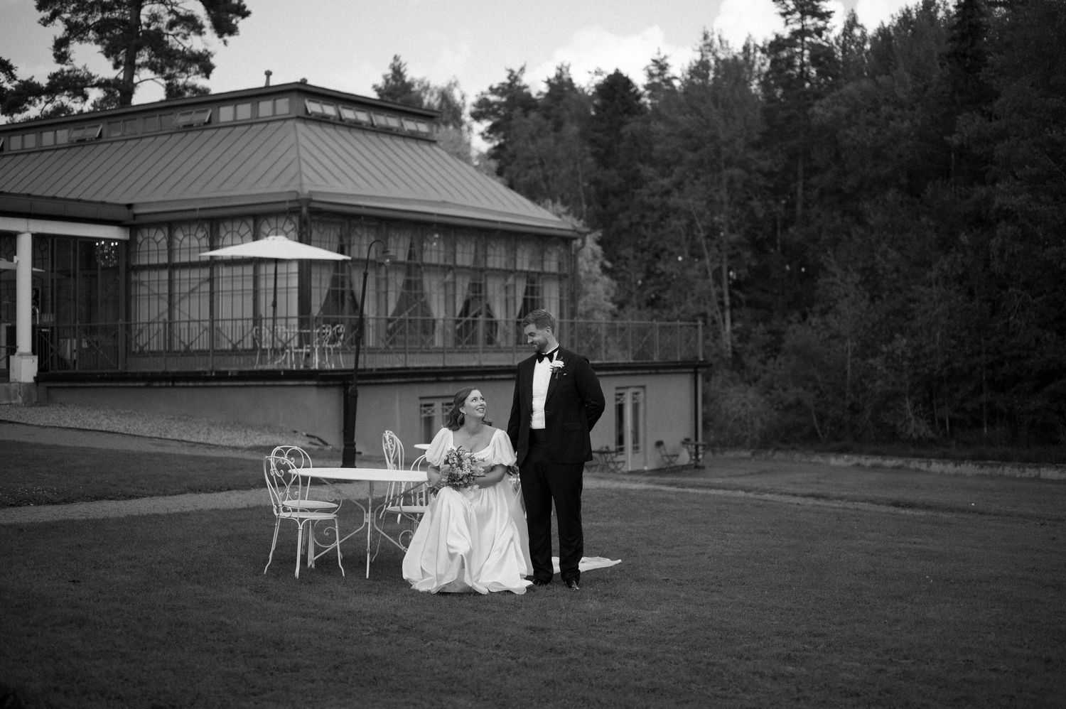 Black and white wedding photo with couple sitting at outdoor furniture near a building.