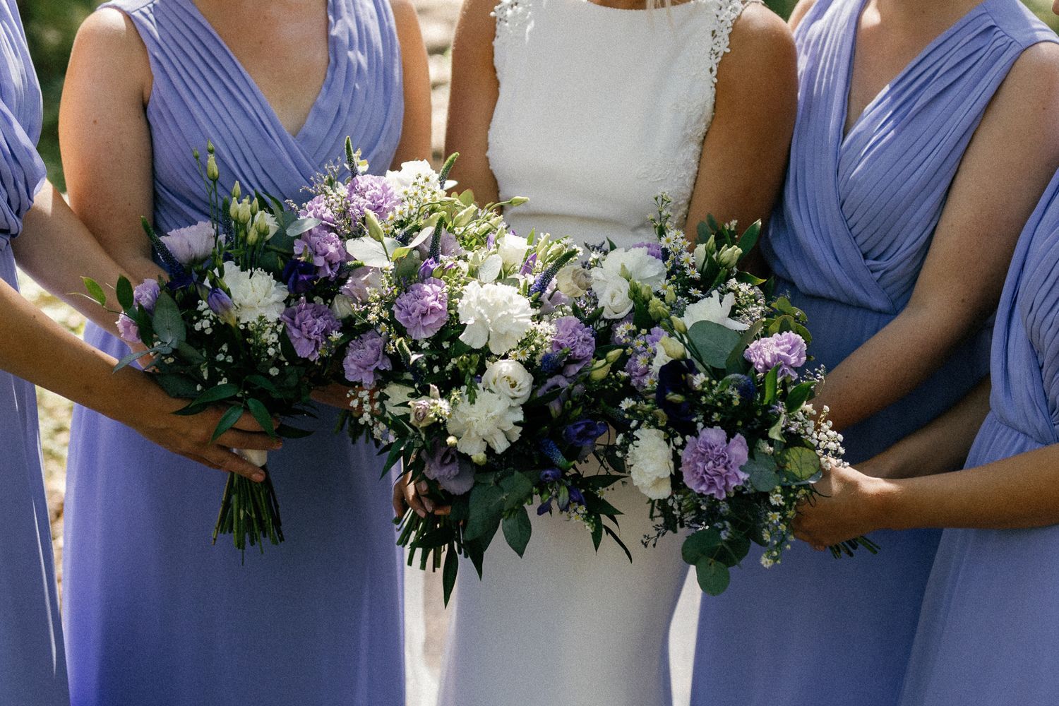 Row of bridesmaids in periwinkle dresses holding white and purple bouquets.
