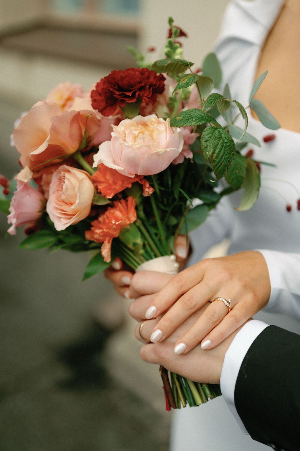 Close-up of wedding bouquet with peach roses and burgundy flowers held by hands with ring.