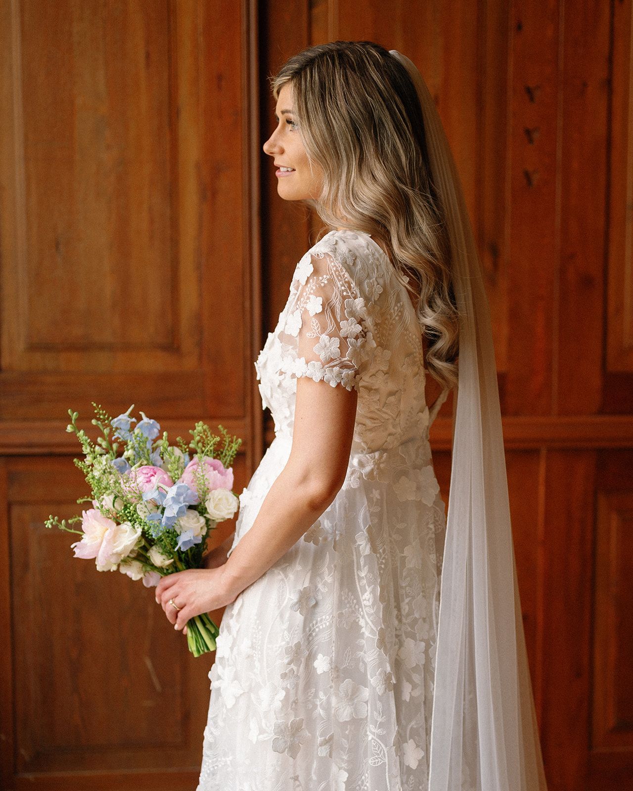 Side view of a bride in a lace dress holding a pastel bouquet against wooden paneling.