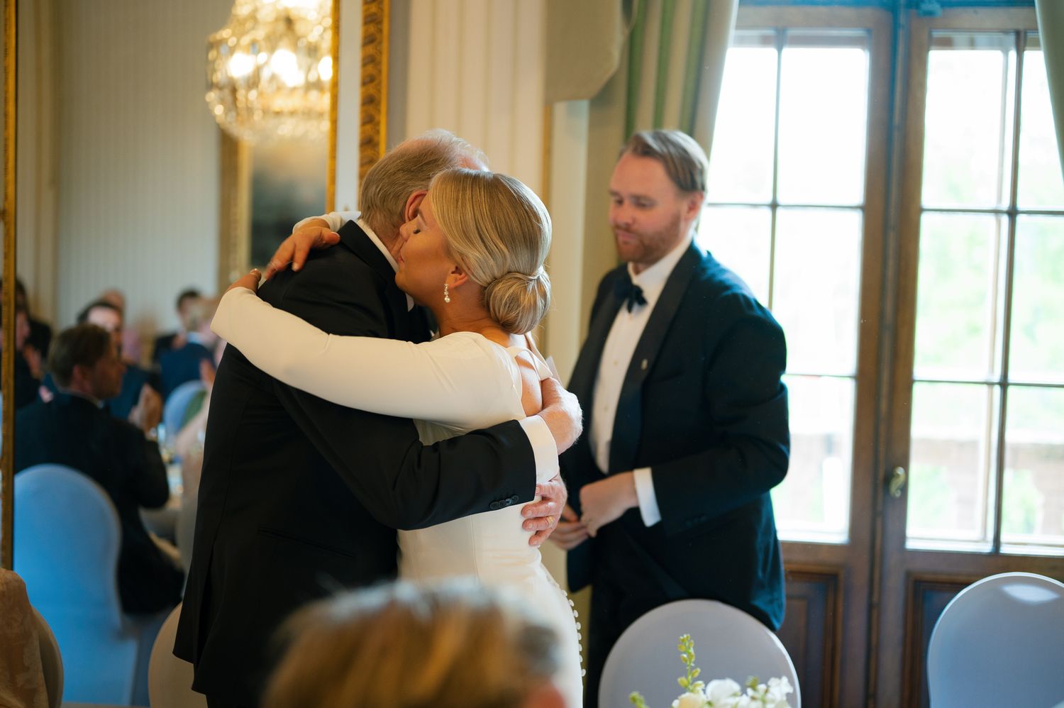 Two people embrace during an elegant event in a formal room with chandelier and blue chairs.