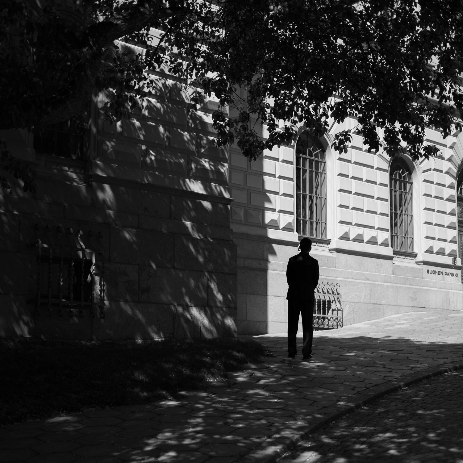 Black and white photo of a lone figure standing on a sunlit sidewalk beneath tree shadows beside a classic building.