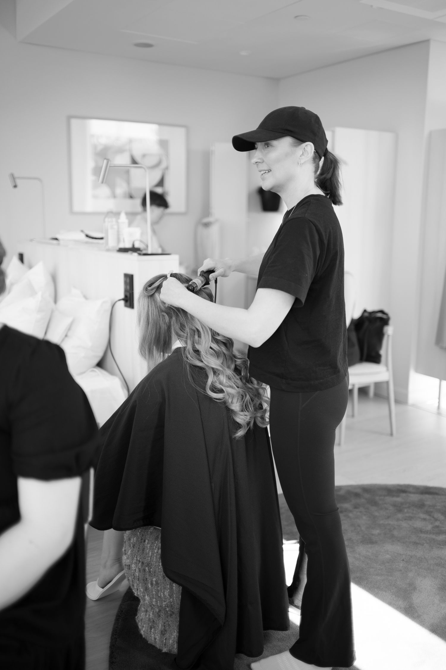 A smiling hairstylist in a black cap and outfit styles a seated client's curly hair in a bright modern salon.