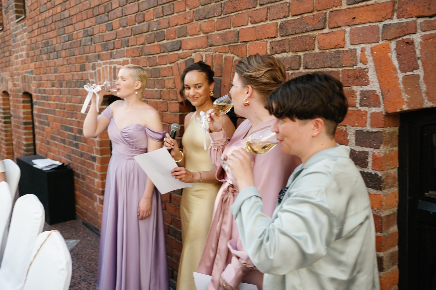Bridesmaids in purple and gold gowns drinking wine at an outdoor wedding reception near a brick wall.