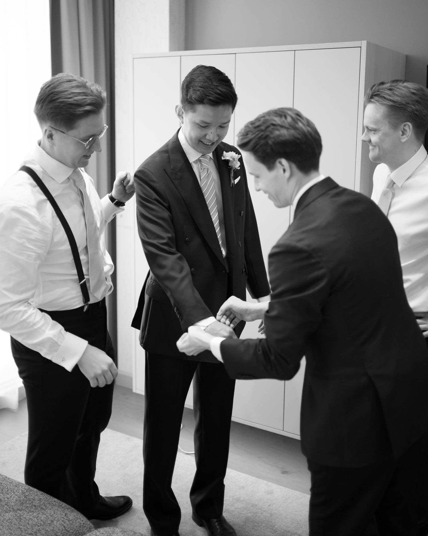 Black and white photo of groomsmen helping groom with cufflinks before wedding ceremony.