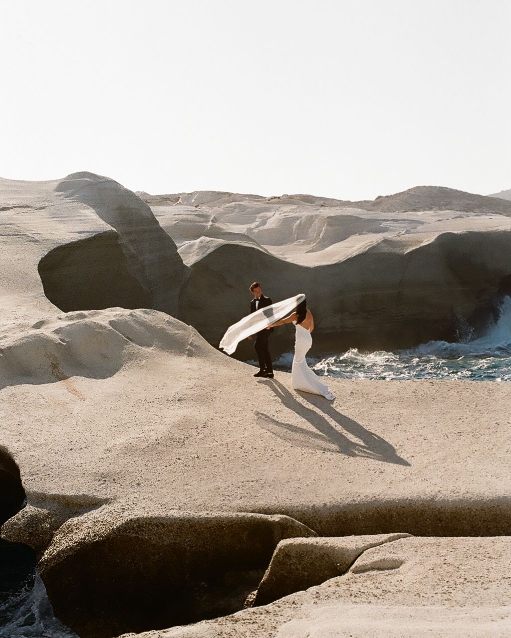 Couple in wedding attire posing on dramatic volcanic rock formations by the sea on a sunny day.
