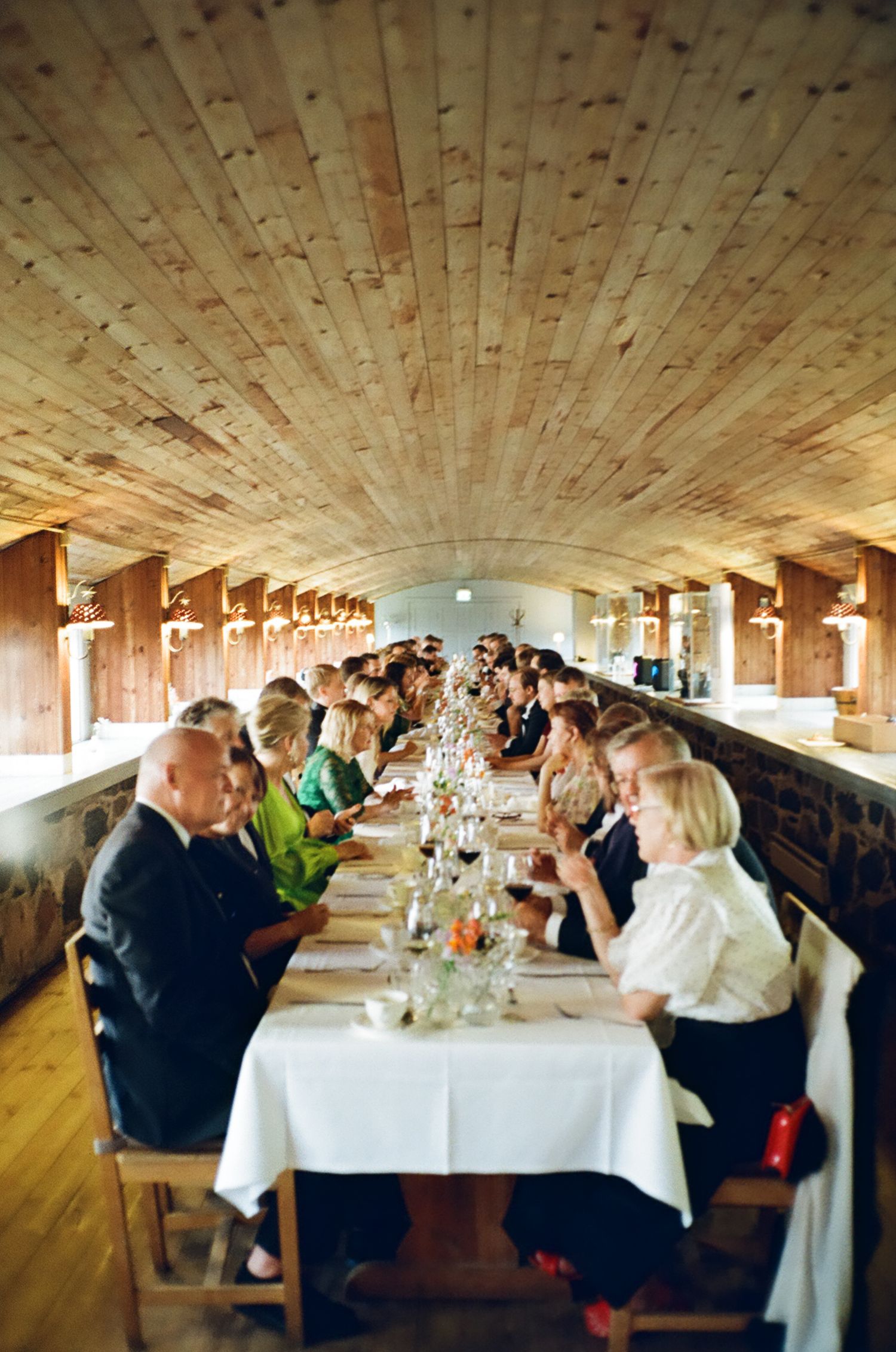 Guests dining at a long banquet table in a rustic wooden hall with arched ceiling during a formal event.