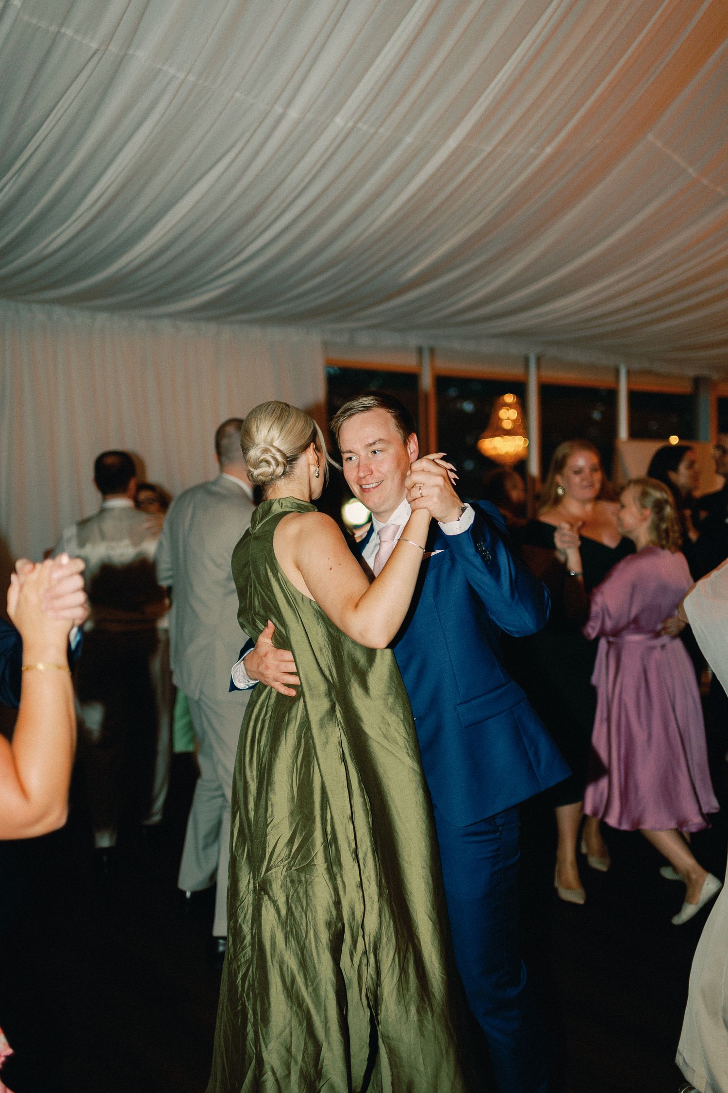 Couple dancing at wedding reception, groom in navy suit, partner in elegant olive green gown, under draped tent ceiling.