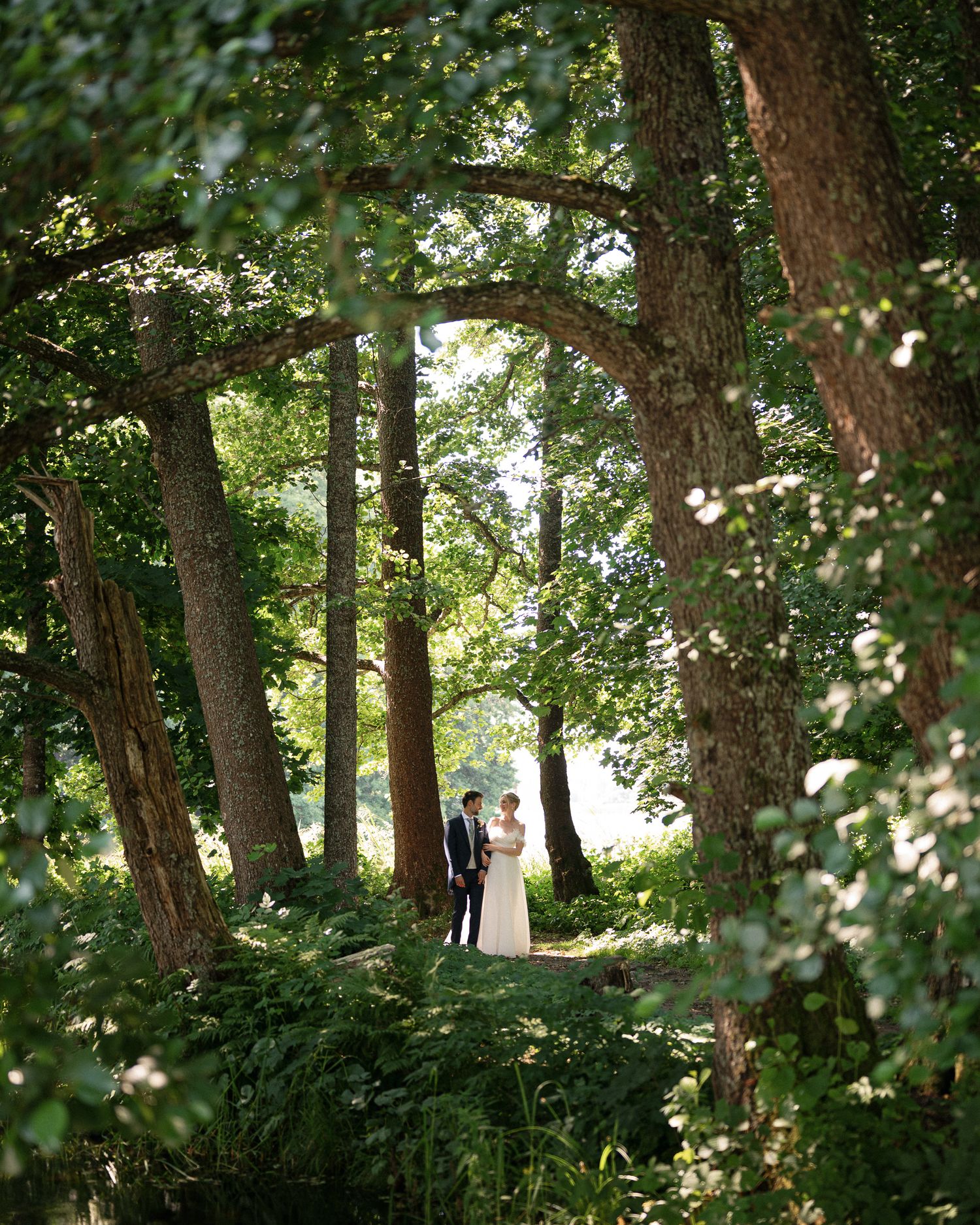A bride and groom stand among towering trees in a lush green forest, framed by natural archways of branches.