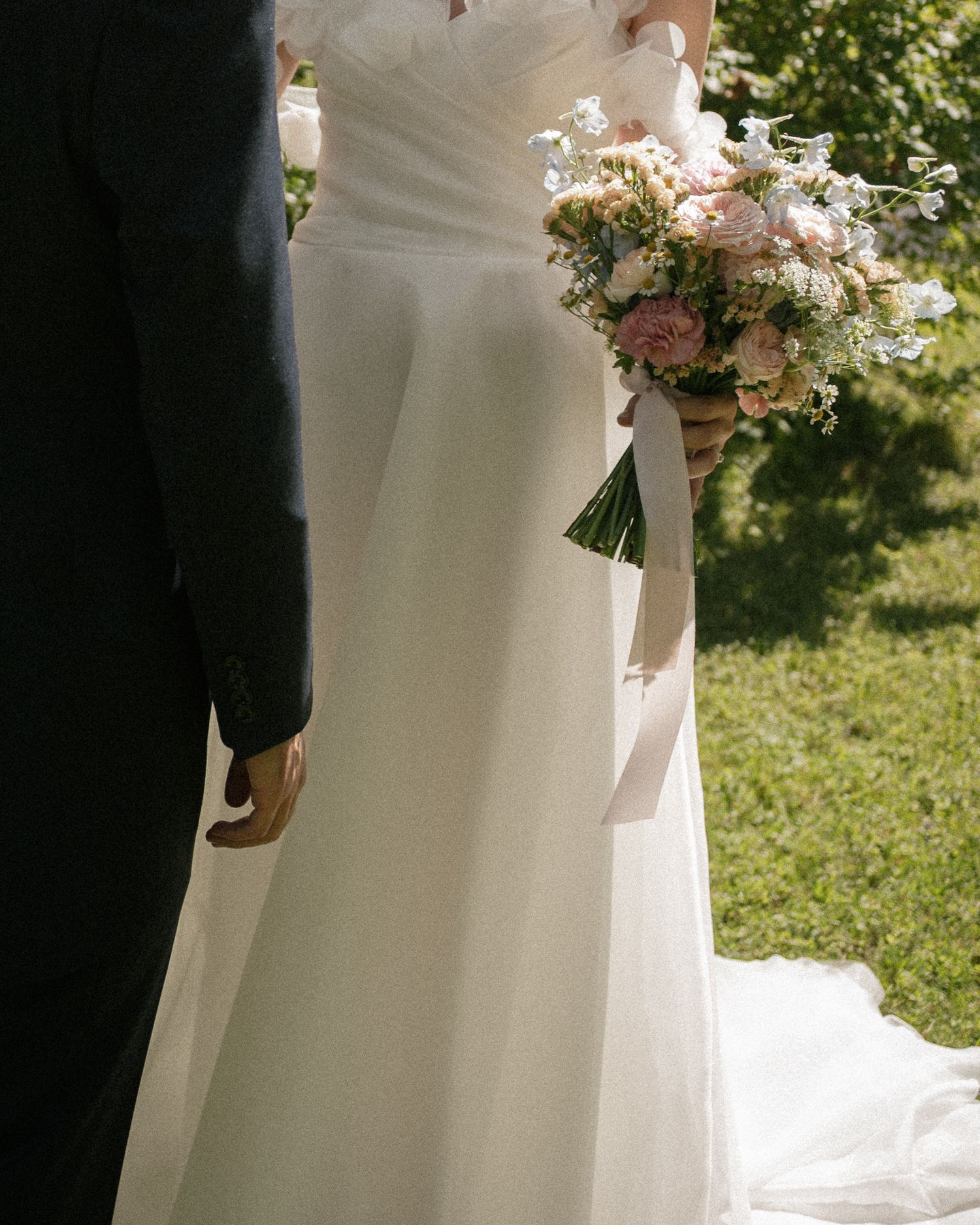 Bride in elegant white wedding gown holding pink floral bouquet stands beside groom in black suit outdoors.