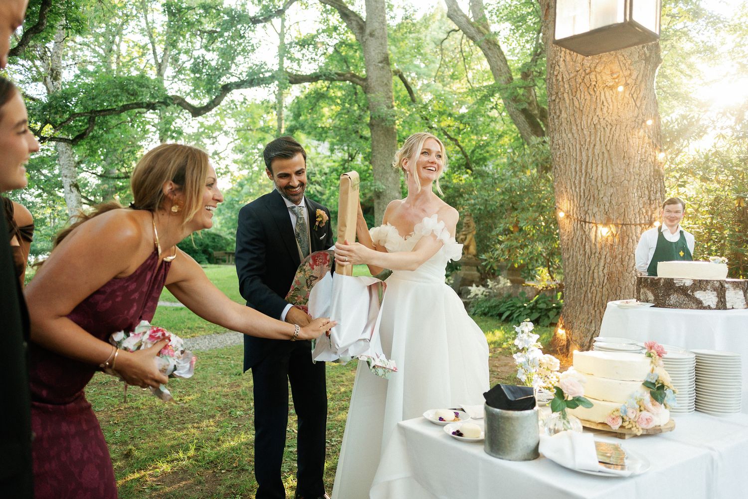 Bride and groom laughing with wedding guests near cake table outdoors under trees with string lights.