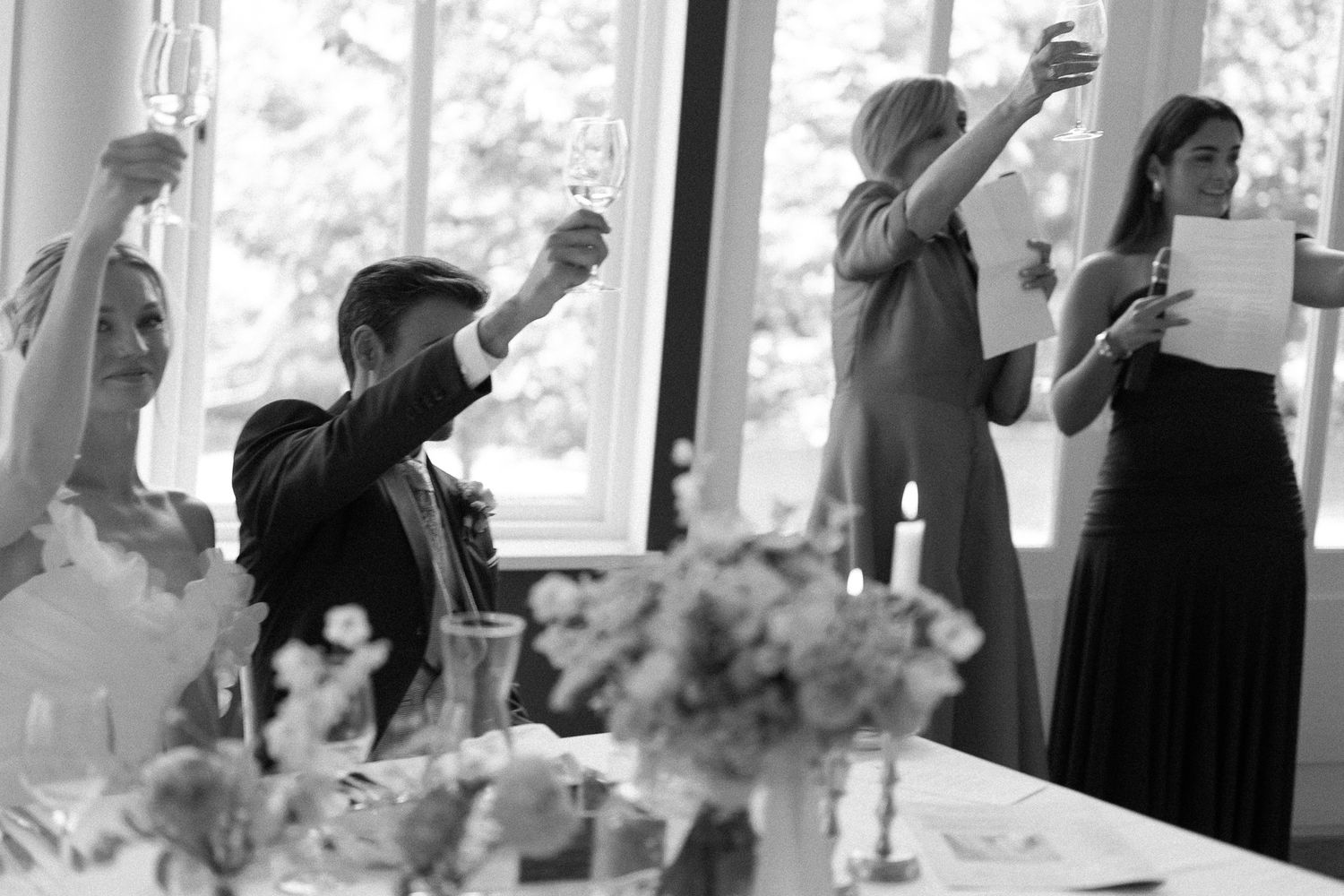 Black and white wedding toast with bride, groom, and guests raising glasses in elegant venue.