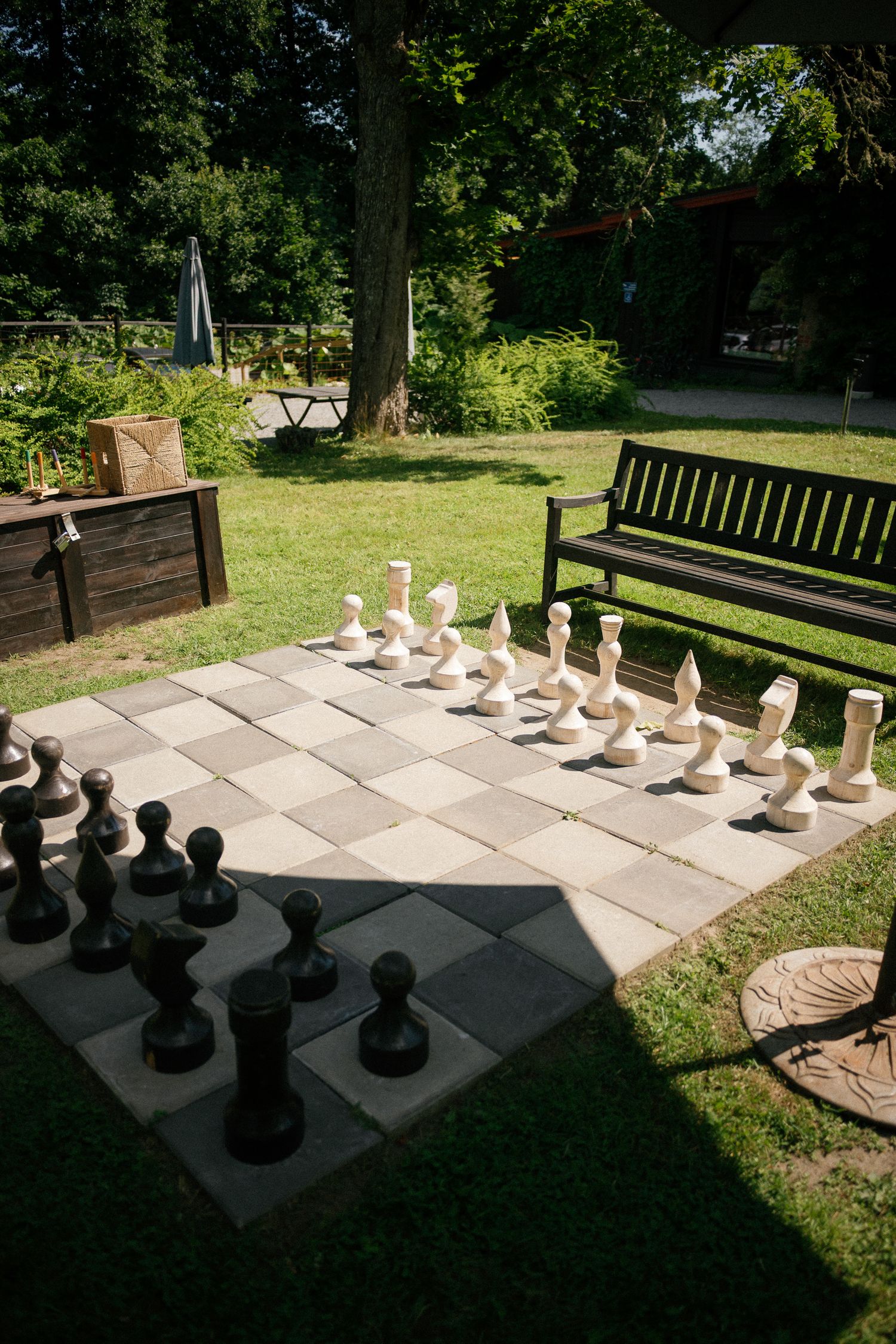 Large outdoor chess set on stone board in sunny park garden with wooden bench and green trees in background.