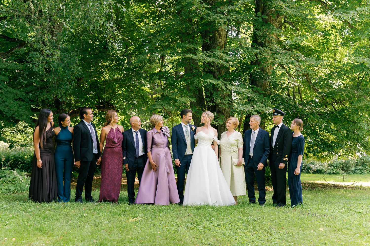 Wedding party poses outdoors on lush green lawn surrounded by tall trees, featuring bride in white gown with family members.