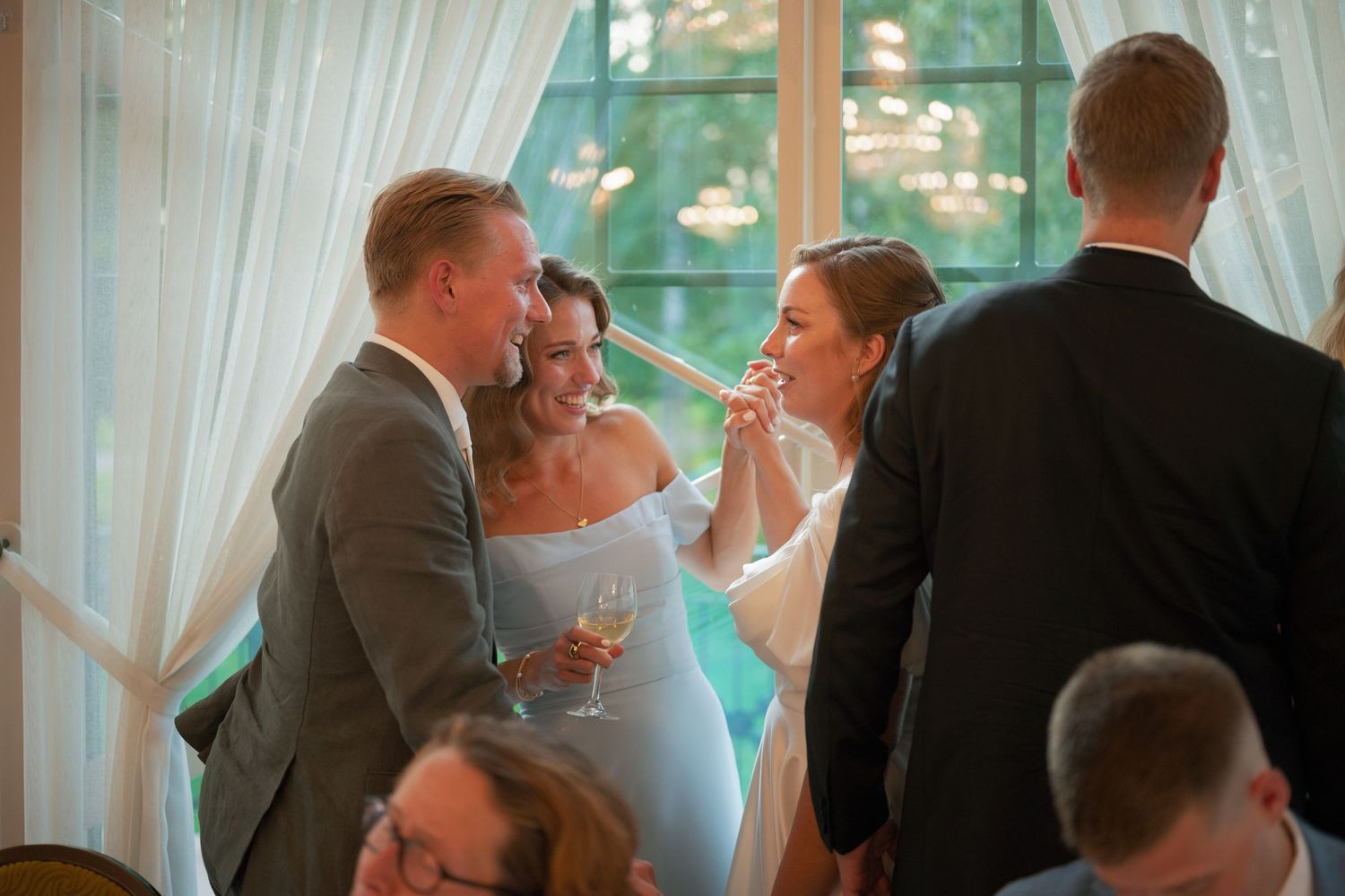 Guests enjoying themselves at a wedding reception in a bright sunlit room with white curtains.