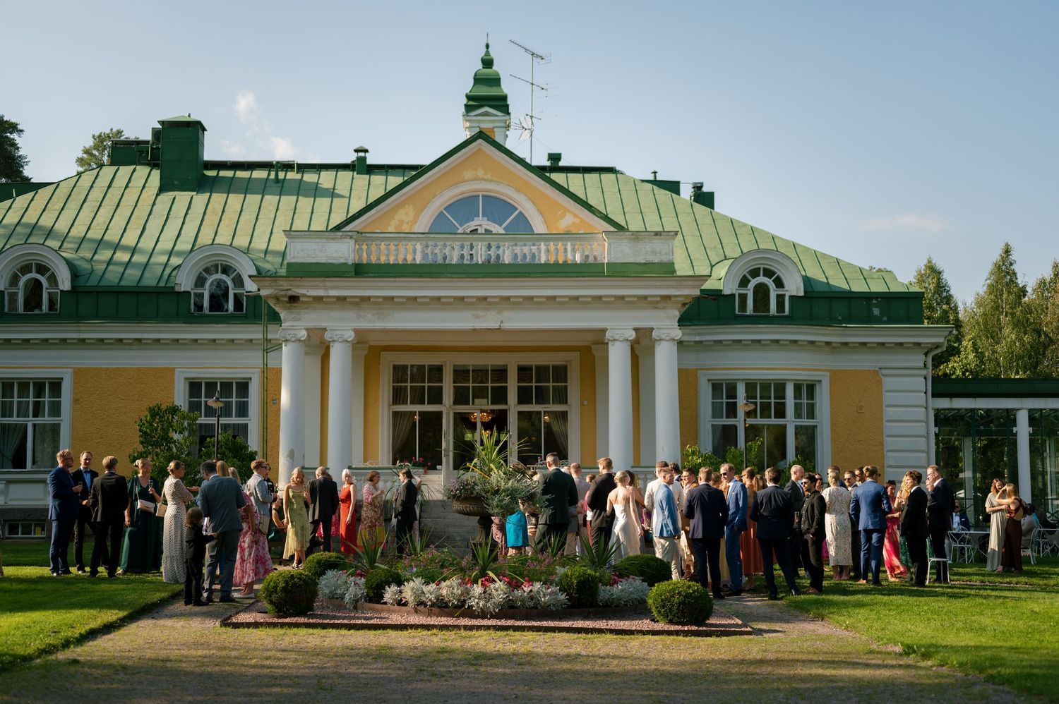 Guests gathered outside a grand yellow mansion with green roof and white columns.