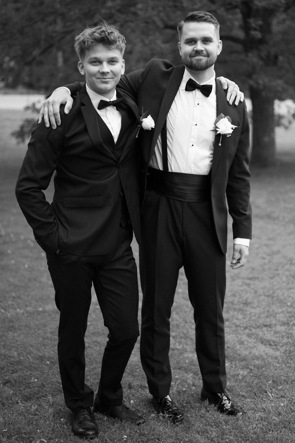 Two men in classic black tuxedos pose together at formal event outdoors.