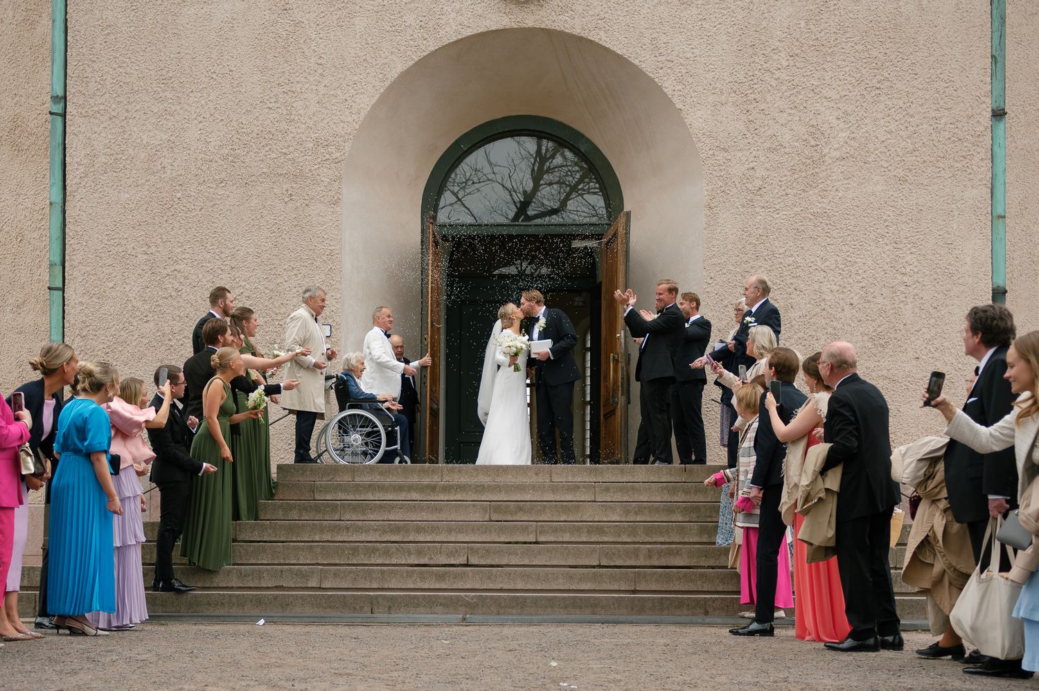 Wedding guests dance in pairs along a stone building exterior during an elegant outdoor reception.
