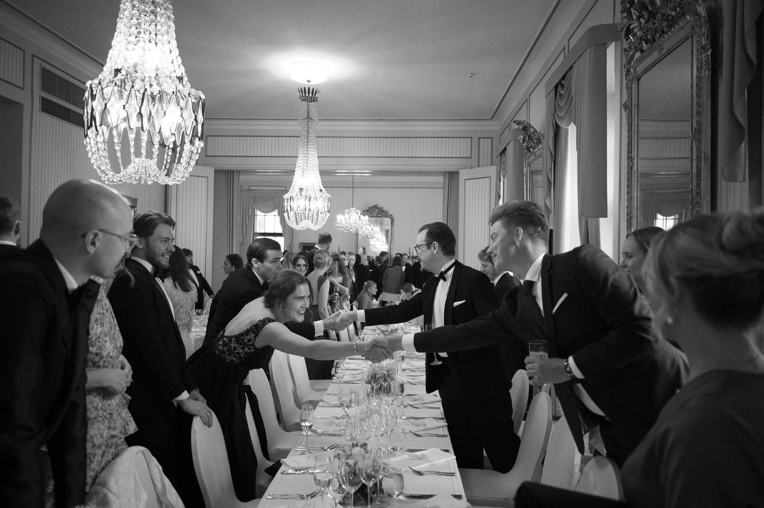 Elegant black and white reception photo showing guests mingling under crystal chandeliers in a formal dining room.