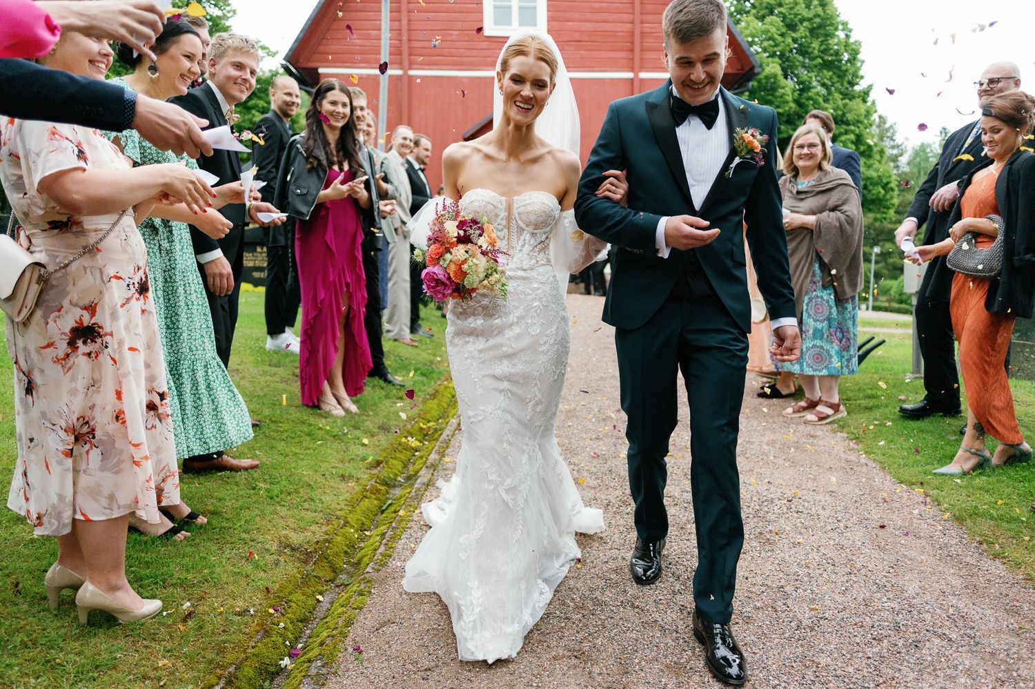 Wedding guests line a dirt path throwing flower petals during a joyful outdoor celebration.