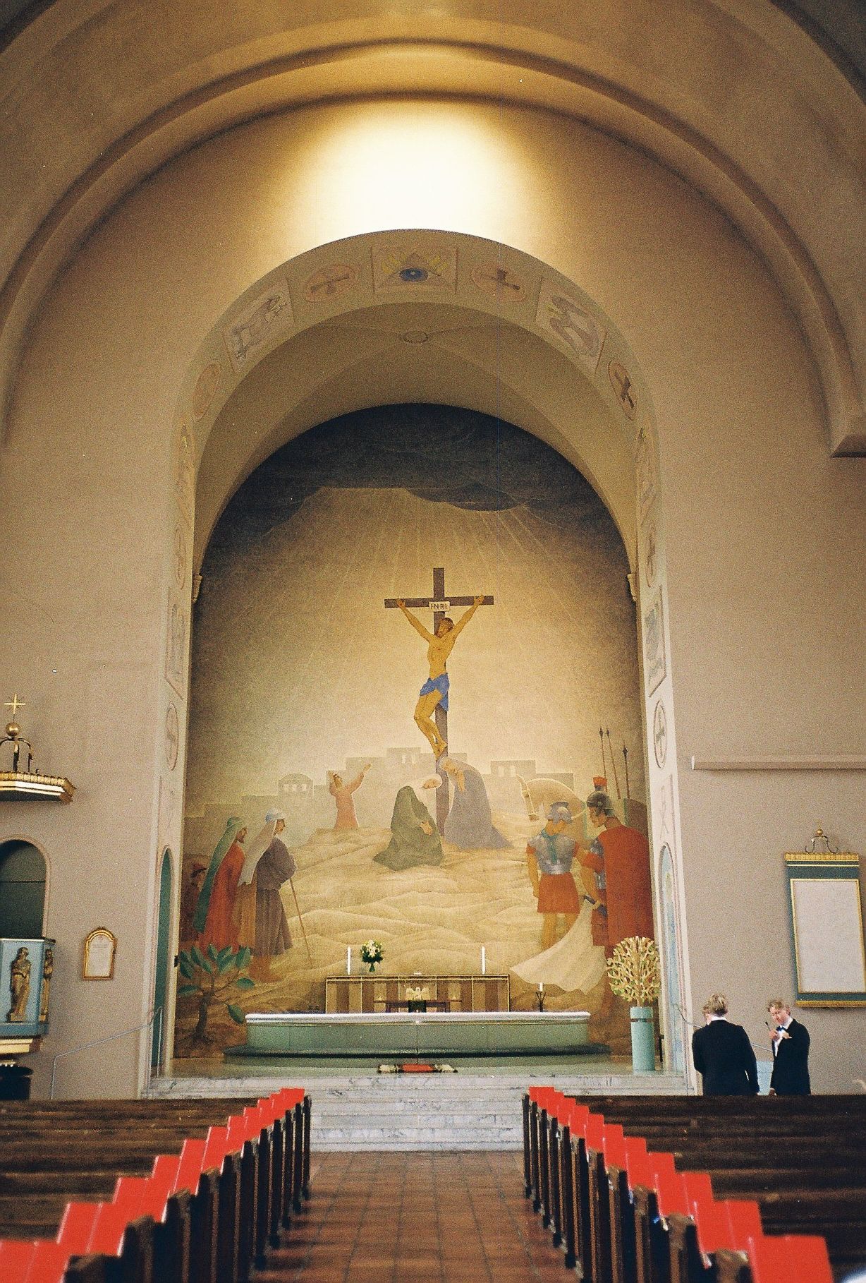 Church interior with religious mural and red pews under arched ceiling.