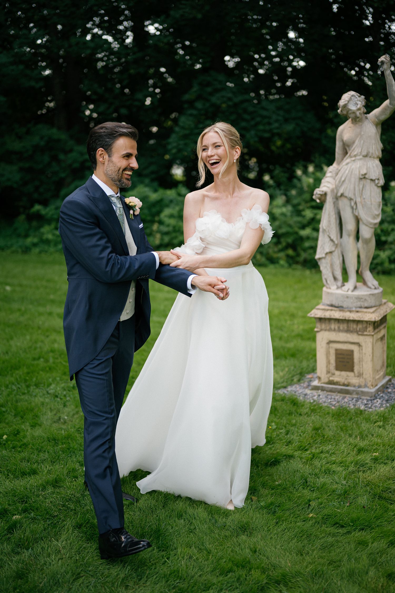 Joyful bride and groom dancing on green lawn beside stone statue, bride in flowing white gown, groom in navy suit.