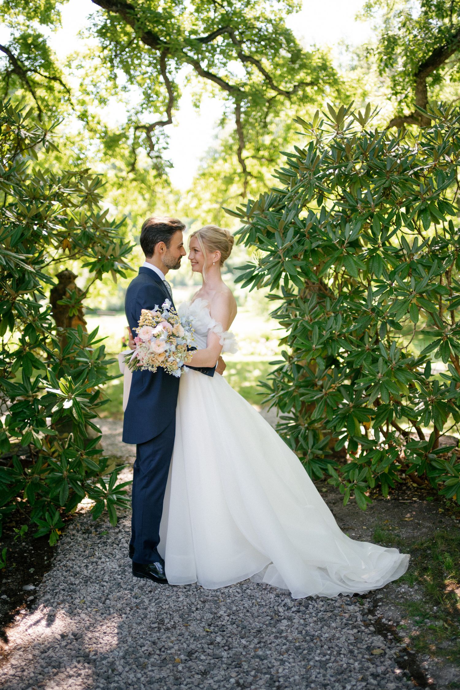 Bride and groom embrace in lush garden path, she in white ballgown holding bouquet, he in navy suit.