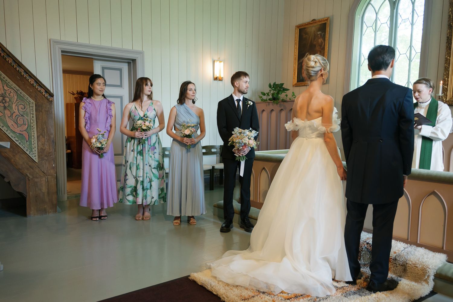 A bride and groom stand at the altar during a wedding ceremony with bridesmaids and officiant in a white chapel.