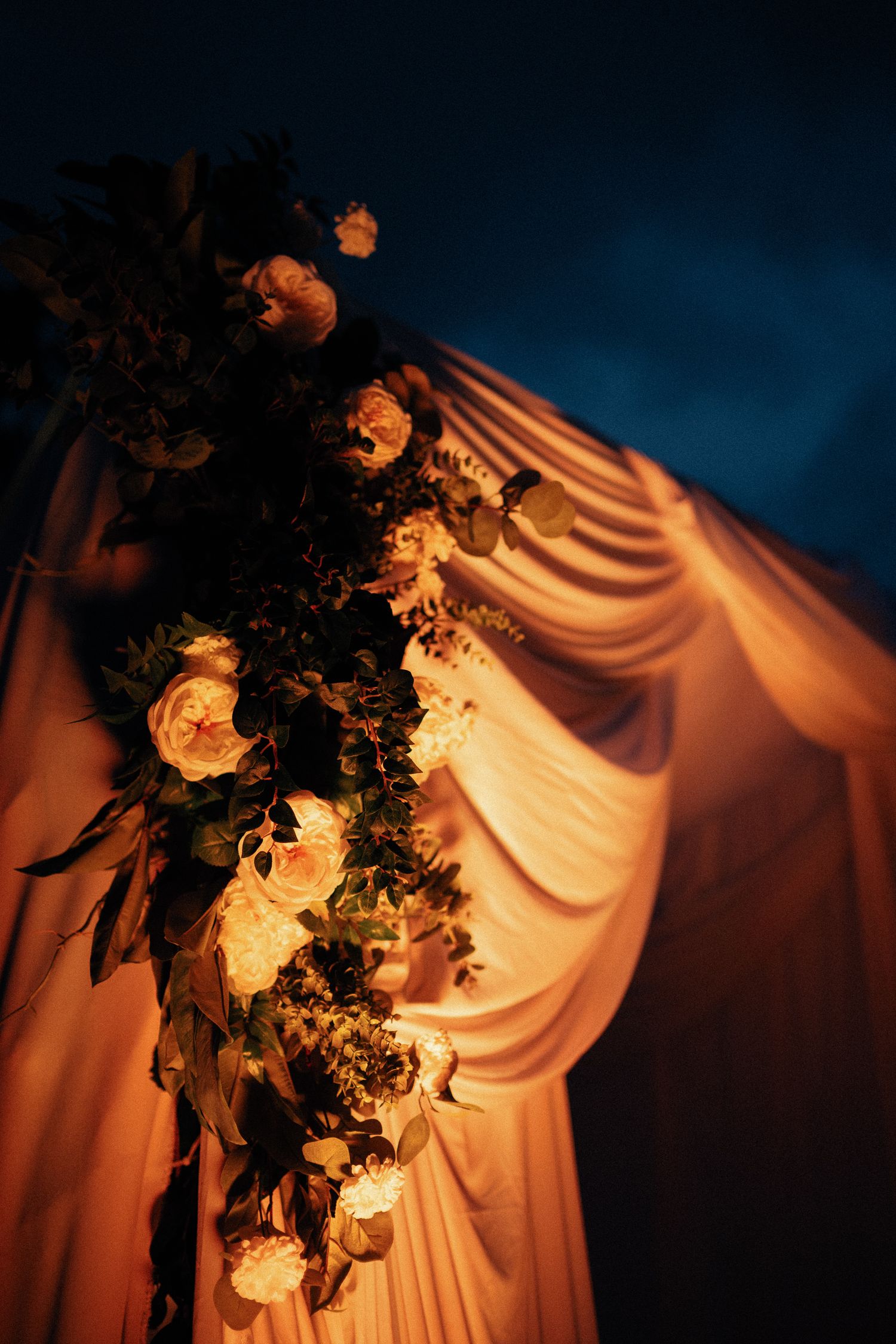 Elegant wedding arch with white roses, greenery, and warm fairy lights against draped fabric at night.