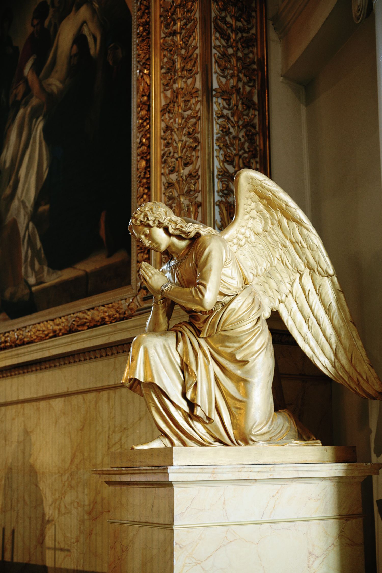 Golden angel statue kneeling in prayer with wings spread, displayed in an ornate church interior with gilded decor.
