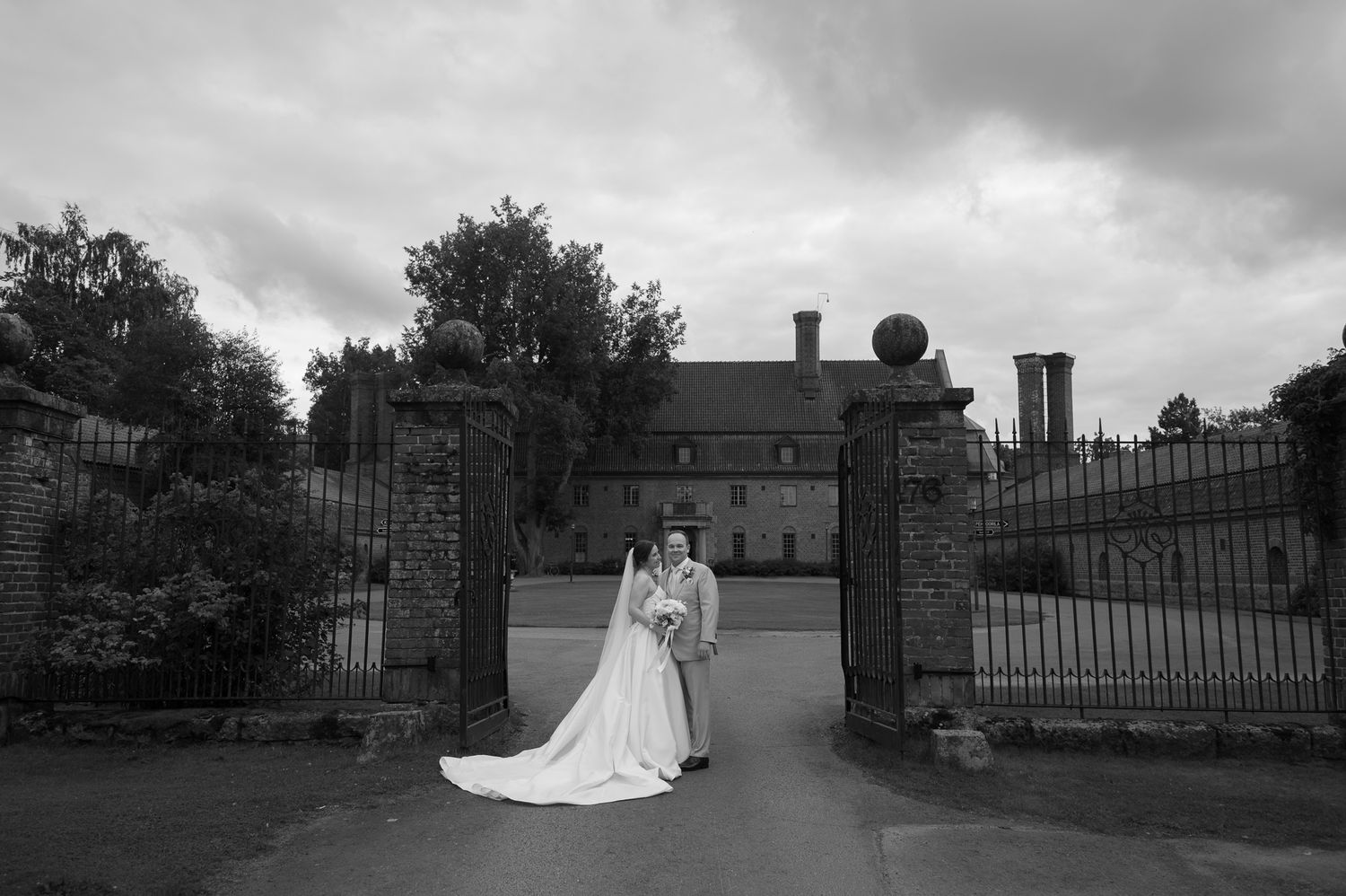 Black and white wedding photo of bride and groom embracing at ornate gate of elegant manor house under dramatic cloudy sky.